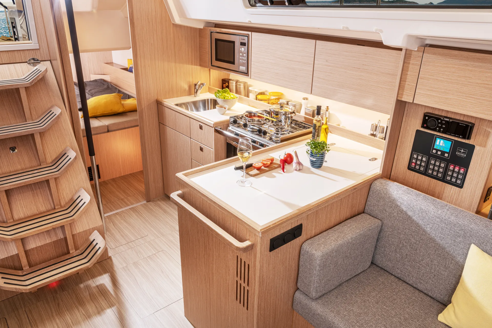 A modern yacht galley aboard the Bavaria C38 features sleek wooden cupboards, a sink, hob, wine glass, and cooking ingredients arranged neatly on the worktop. A grey sofa and control panel are nearby, with stairs leading to another area and a bed visible in the background.