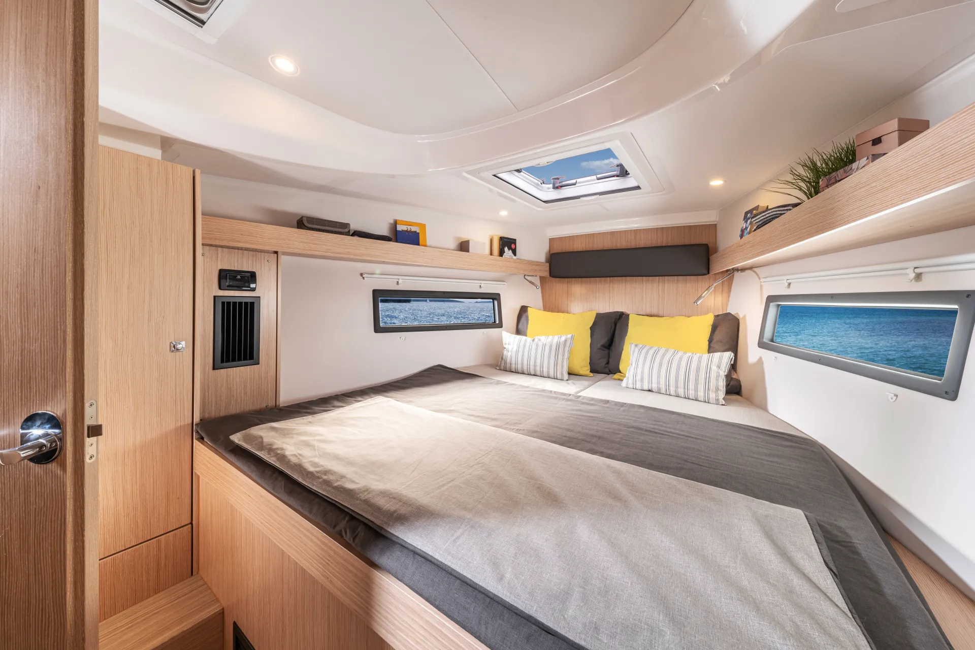 Modern boat cabin with wood accents, a large bed with grey and yellow pillows, windows on both sides showing the sea, and a ceiling skylight letting in natural light. Thoughtfully designed like the interior of the Bavaria C38, shelves hold books and small decorations.