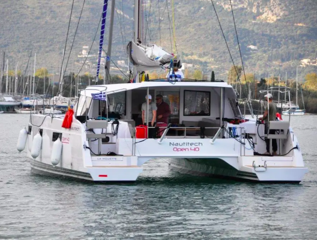A catamaran docked at a marina with mountains in the background. Two people are visible on the boat, which has a green dinghy hoisted at the rear.