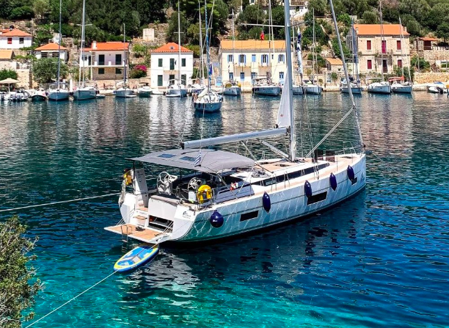 A yacht with a green and white hull moored in a calm harbor filled with several boats, set against a background of small colorful houses and lush greenery on a sunny day.