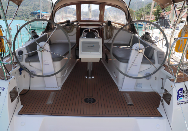 A view from the stern of a sailboat showing dual steering wheels, a binnacle with a chart plotter, and teak decking. The harbor and green hills are visible in the background. This