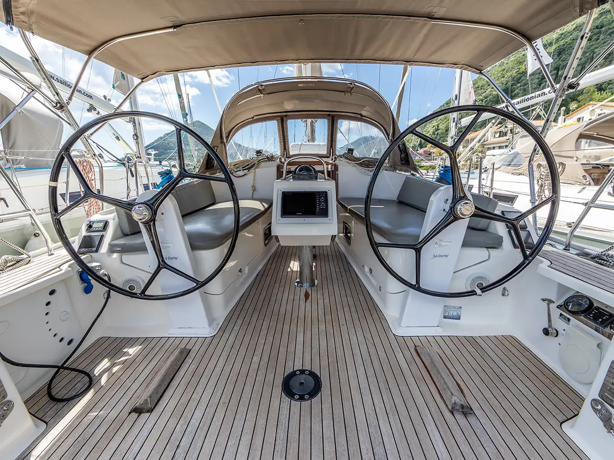 A view of a Bavaria 37 sailing yacht's cockpit, featuring dual steering wheels, a central navigation console, and cushioned seating areas on both sides. The cockpit is under a canopy with a clear view of the marina and surrounding boats in the background.