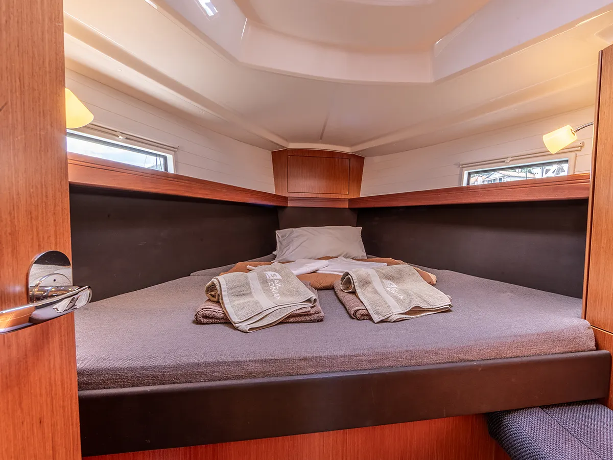 The image shows the interior forepeak cabin of a Bavaria 37 with a neatly made bed. The bed has gray bedding with two folded towels placed on top. The cabin features wooden paneling, two small windows on either side, and ambient lighting, creating a cozy and inviting atmosphere.