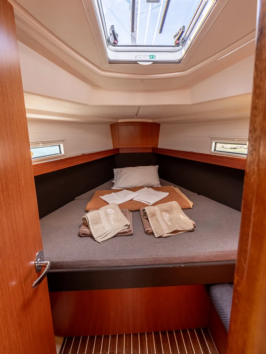 A cozy Bavaria 37 forepeak cabin with a neatly made bed featuring gray bedding, two white pillows, and folded towels at the foot. The cabin has wooden walls and flooring, with windows on both sides and a ceiling hatch letting in natural light.