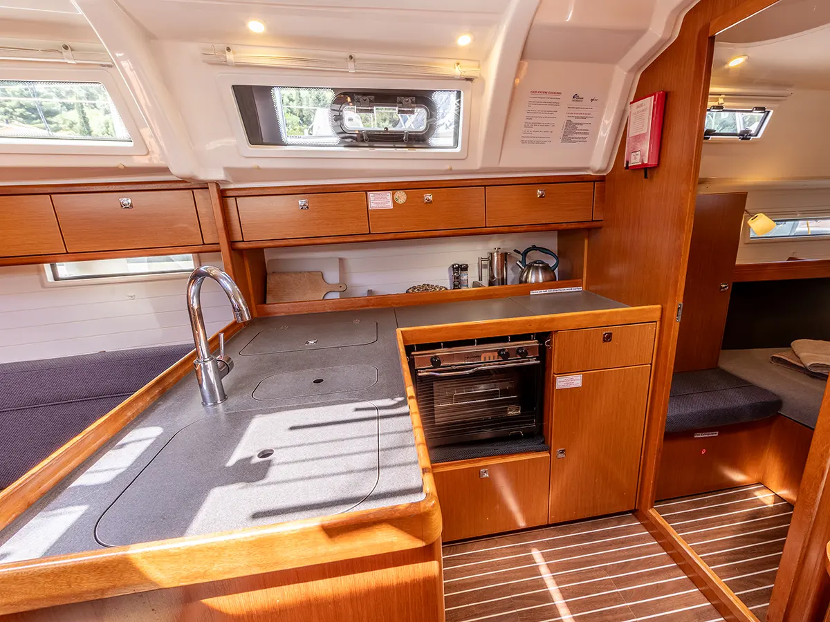 A yacht galley with wood cabinetry, a large sink with a single-handle faucet, a built-in oven, and a stovetop. The space is well-lit with natural light coming through the windows and a skylight. Adjacent to the galley is the dining area and a cozy sleeping nook.