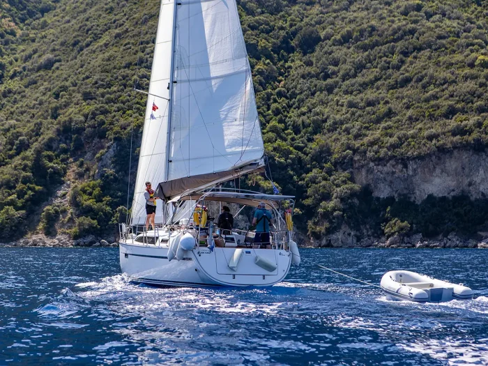 A white Bavaria 37 sailing yacht with several people aboard sails near a rocky, forested coastline. The boat tows a small inflatable dinghy and the water is calm with gentle waves under a clear blue sky. The scene exudes a tranquil, leisurely maritime adventure.