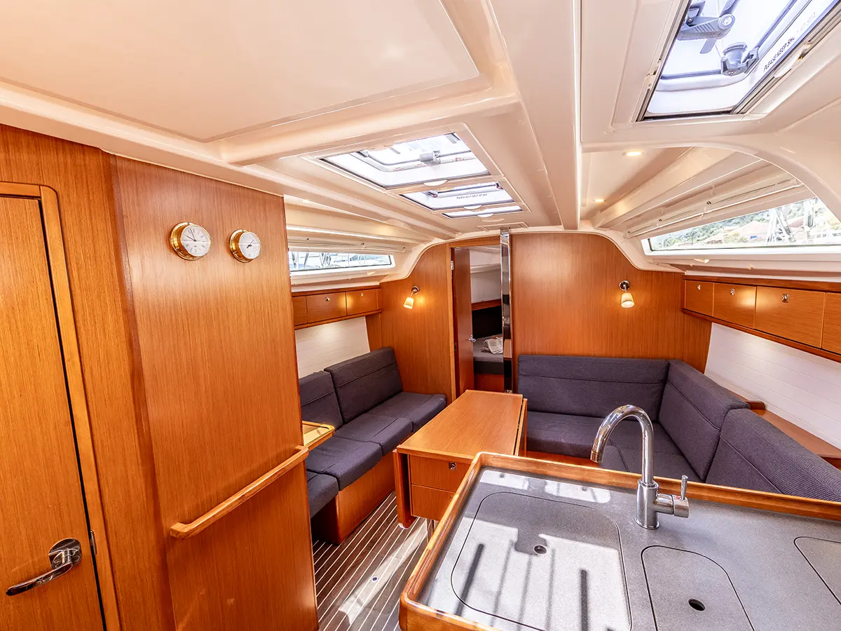 The image shows the interior of a modern sailboat. It features wooden furnishings, with two gray upholstered seating areas along the sides and a dining table in the middle. The foreground has a small kitchen sink, and sunlight streams in through overhead windows.