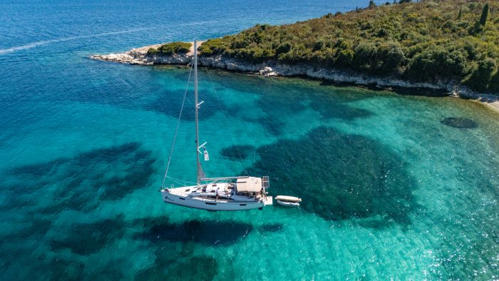 A sailboat is expertly anchored in a serene, turquoise bay surrounded by lush greenery on anchor. The clear water reveals the rocky seabed beneath, creating beautiful patterns and shades. A charter skipper's dinghy is tied to the boat, floating gently on the surface.