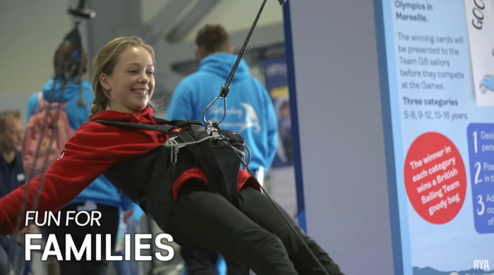 A smiling girl in a red jacket enjoys a sailing simulator at the RYA Dinghy Show. She is seated in a harness, leaning back and holding onto ropes. The background includes a blurred poster and people in blue clothing. Text on the image reads, "Fun for Families.