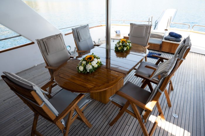A wooden yacht deck, perfect for dining on the sun deck, features a table with two floral centerpieces surrounded by six cushioned chairs. It overlooks calm blue water as the sun casts soft shadows across the polished wood surface.