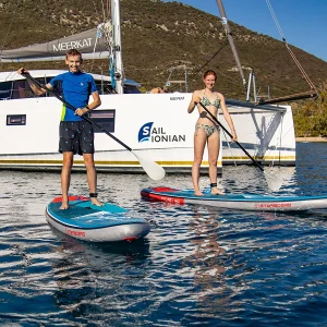 A man and a woman enjoy paddleboarding on calm blue waters near the elegant yacht "MEERKAT," part of the esteemed SAIL IONIAN fleet. They smile amidst lush green hills under a clear blue sky, perfectly capturing the serene charm of an Ionian adventure.