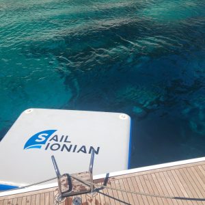 A serene view of clear turquoise water near rocky cliffs unfolds from the deck of a boat emblazoned with "Sail Ionian." The landscape, lush with greenery on the cliffs above, hints at hidden islands waiting to be explored.