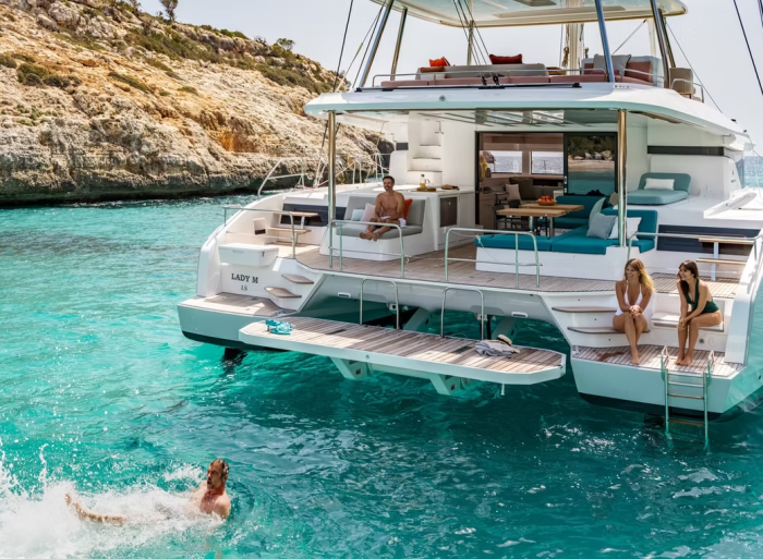 Lagoon 55 catamaran A luxury catamaran yacht anchored in clear turquoise water near rocky shores. Two people sit at the back while another relaxes inside. A person is swimming near the yacht, enjoying a sunny day.