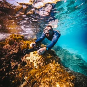 A person wearing goggles swims underwater near a coral reef, holding a Subnado Underwater Scooter. They are gliding effortlessly. Sunlight filters through the clear blue water, illuminating vibrant corals and reflecting above.