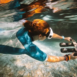 A person wearing a wetsuit, goggles, and wristbands uses a Subnado underwater scooter to glide above a rocky seabed in clear blue water. Sunlight reflects off the surface above them.