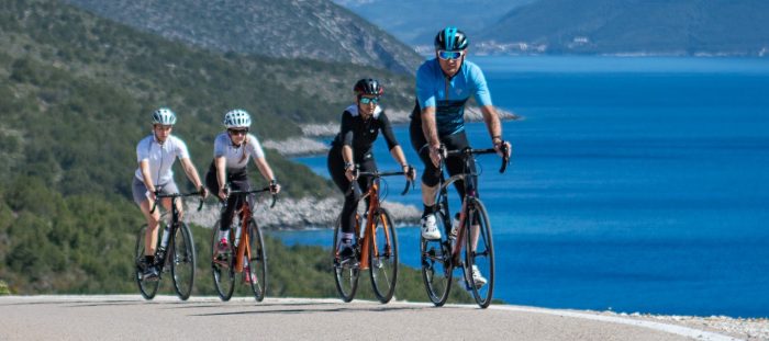 Four cyclists ride on a paved road along a scenic coastline, enjoying biking Lefkada with blue water and green hills in the background under a clear sky.