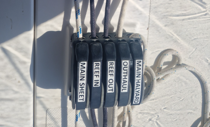Close-up of five labeled sailing rope clutches for Furling Mainsail control: "MAIN SHEET," "REEF IN," "REEF OUT," "OUTHAUL," and "MAIN HALYARD," each holding a different colored rope against a white background.