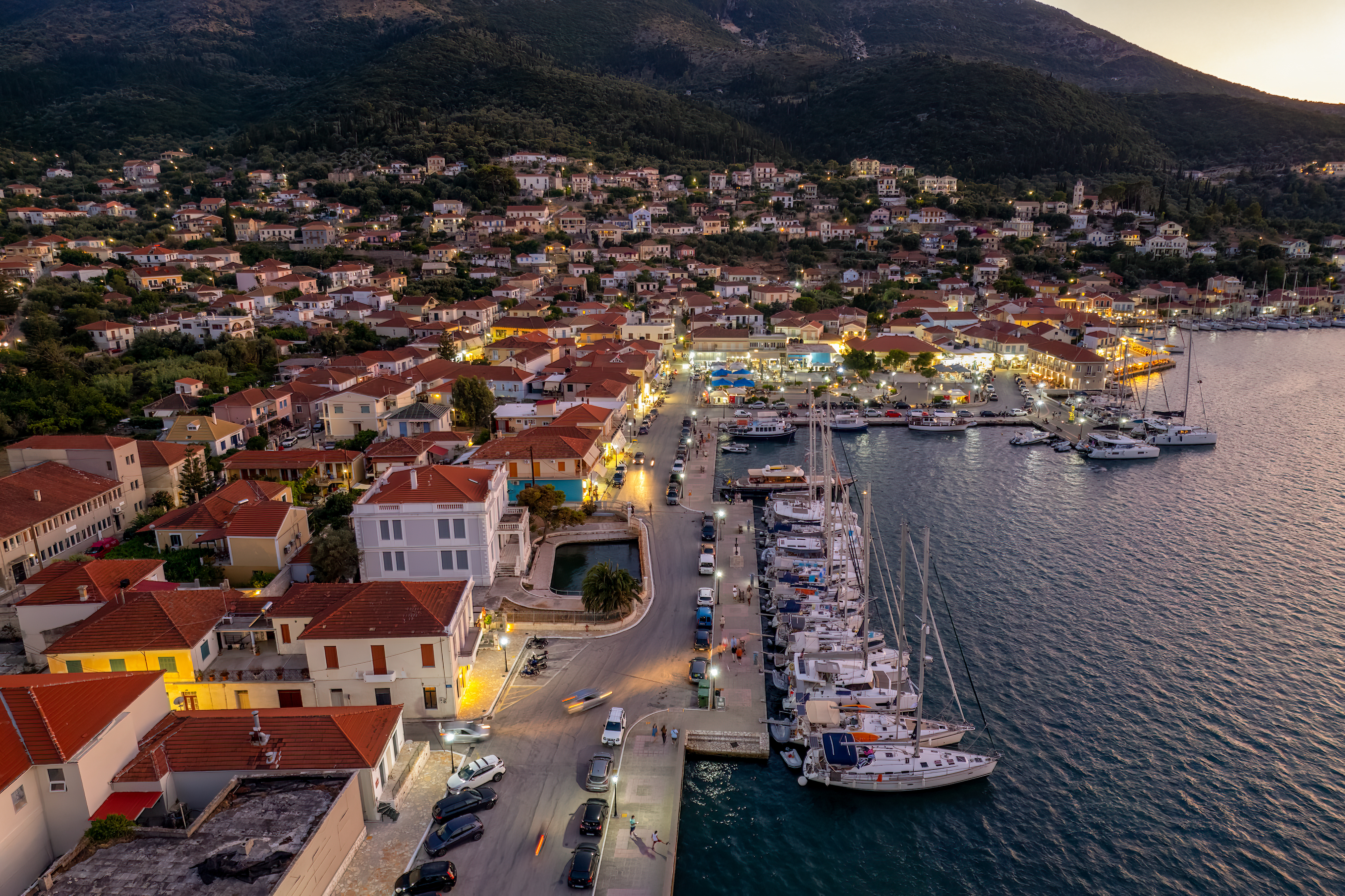 Vathi Ithaca is part of a two eek sailing itinerary from Sail Ionian. Aerial view of a coastal town at dusk, just two weeks south, featuring red-roofed buildings, a marina with moored sailing boats, and hills in the background. The town glows under streetlights as evening sets in.