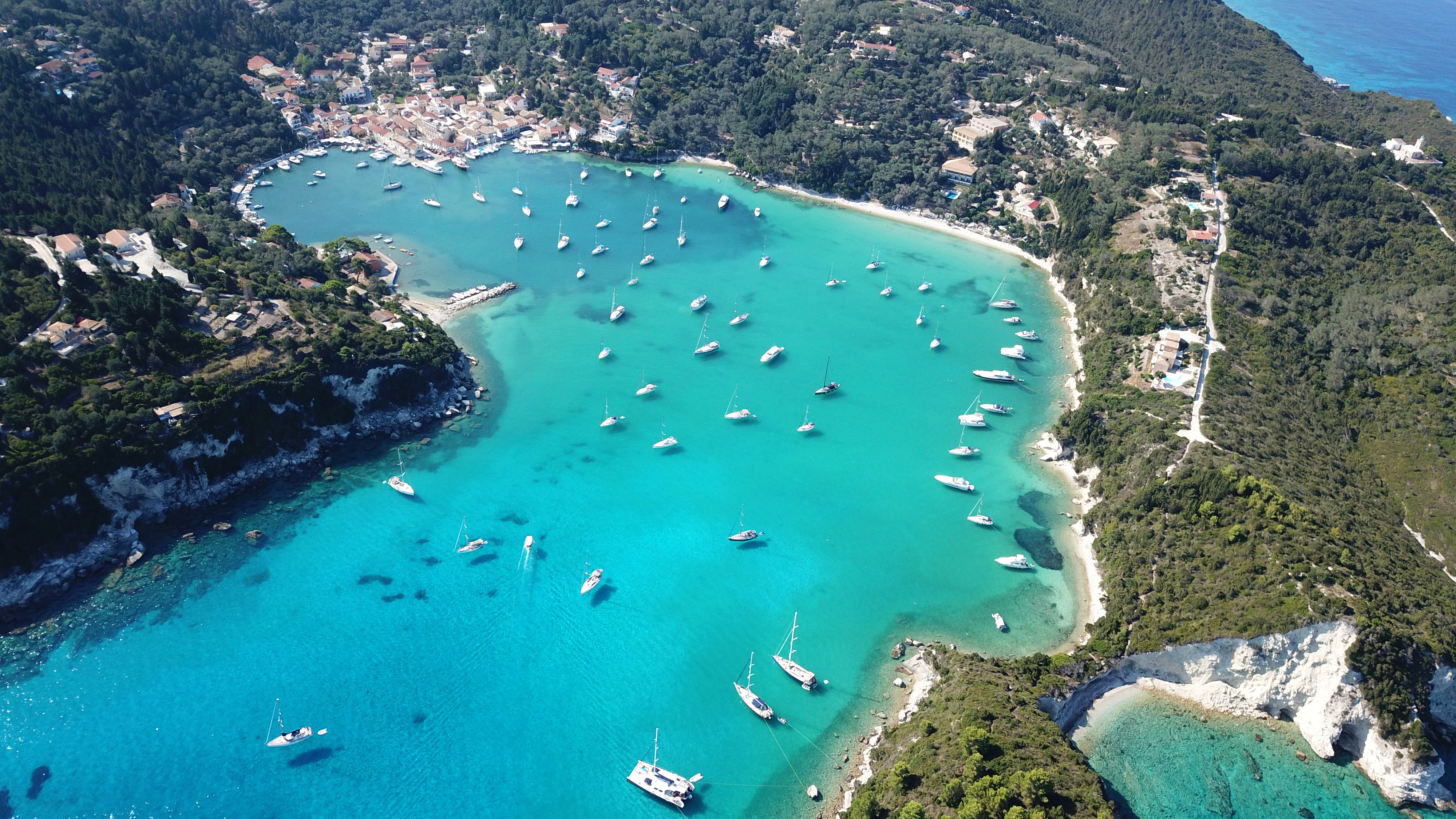 Aerial view of Lakka on Paxos a turquoise bay with numerous white boats anchored in clear water, surrounded by lush green hills and a small coastal village—an idyllic escape just two weeks south of the bustling city.