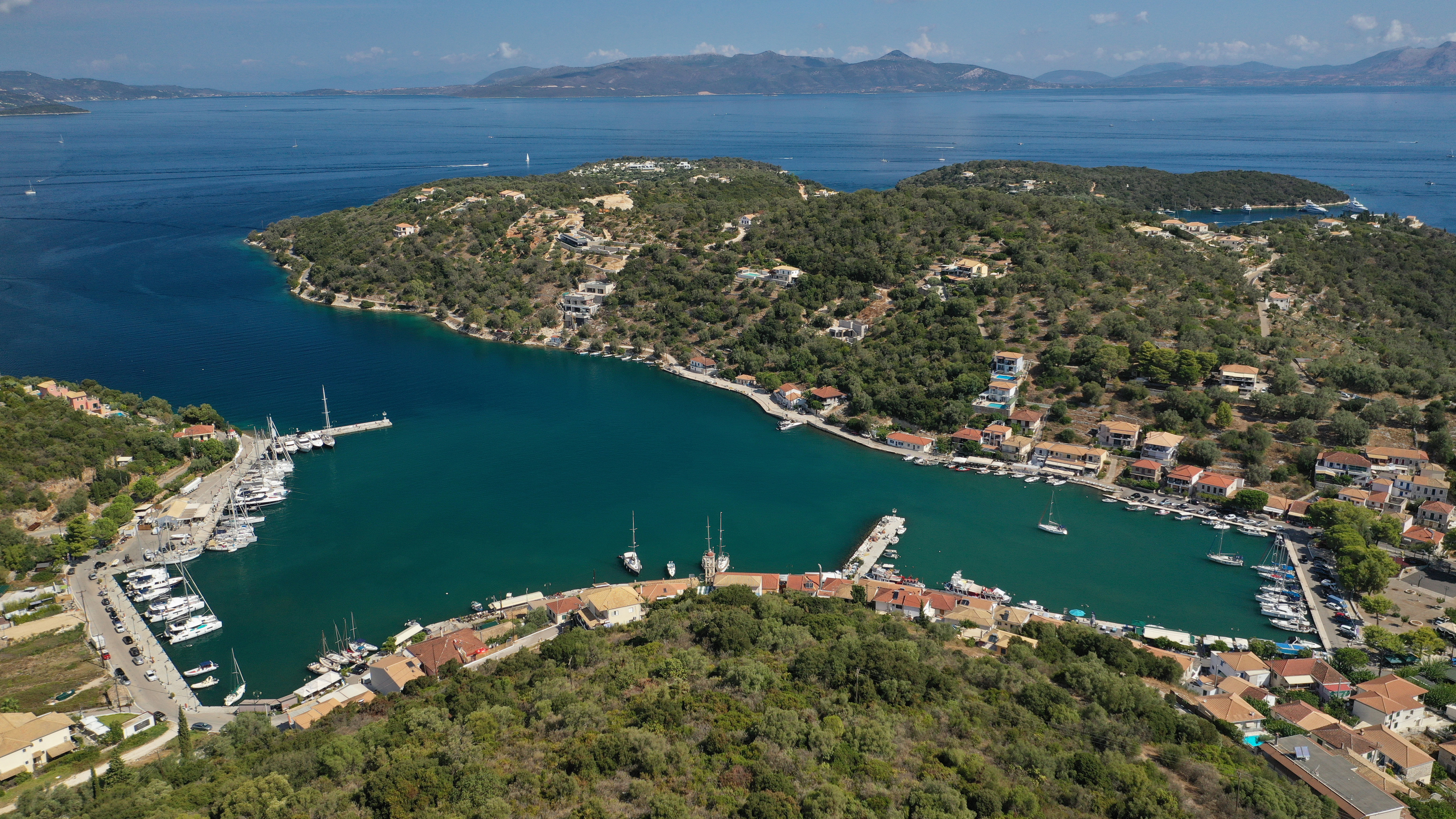 Aerial view of a Little Vathi, Megaisi on Sail Ionian Lively Nightlife route plan, a small coastal village with boats moored along a marina—an ideal spot for quiet escapes—surrounded by green hills and clear blue water, with distant islands and mountains completing the route plan.