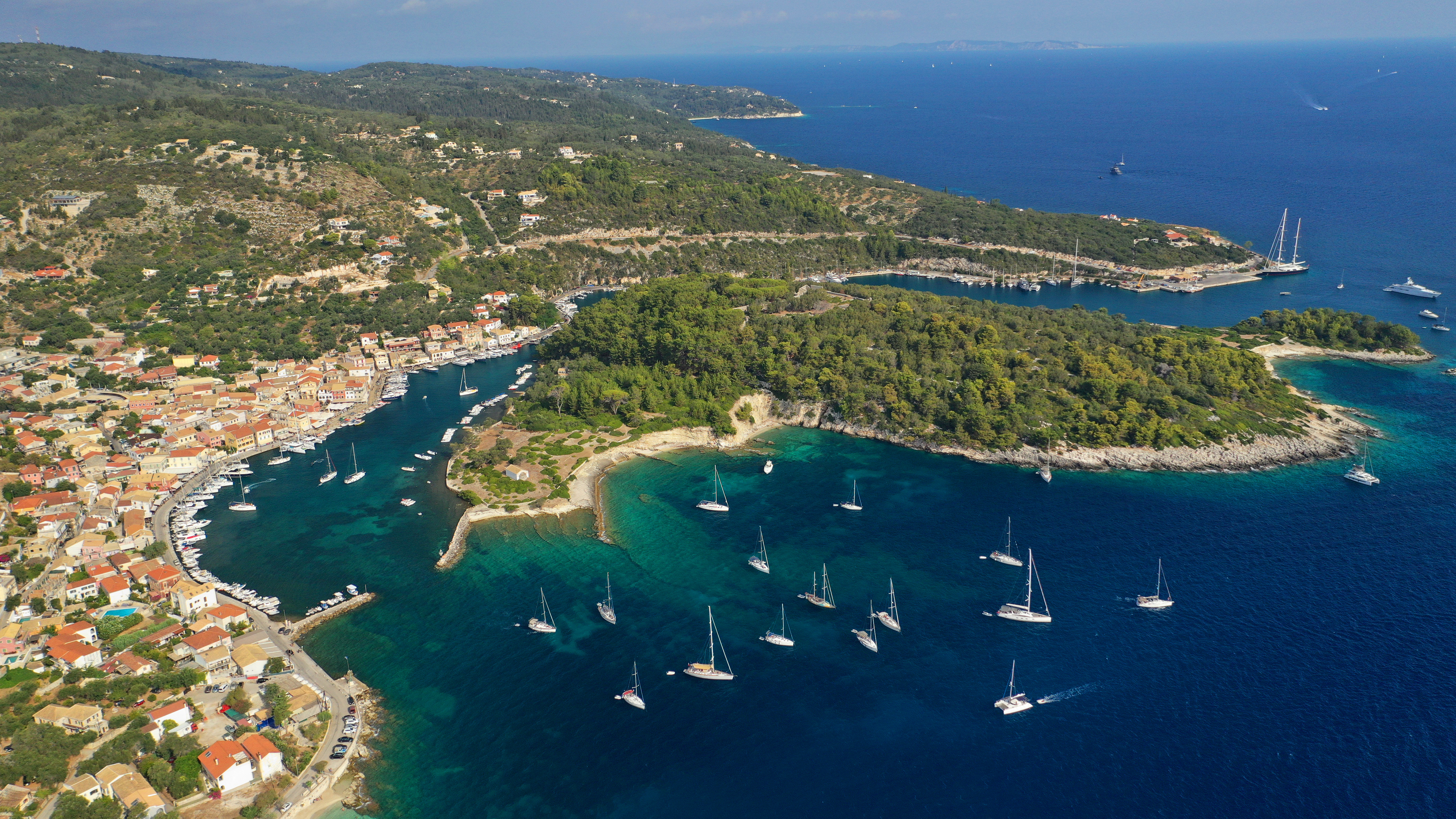 Aerial view of a coastal town with red-roofed buildings, a harbour filled with sailboats, lush green hills, and clear blue sea—your perfect escape fortnight south on a sunny day.