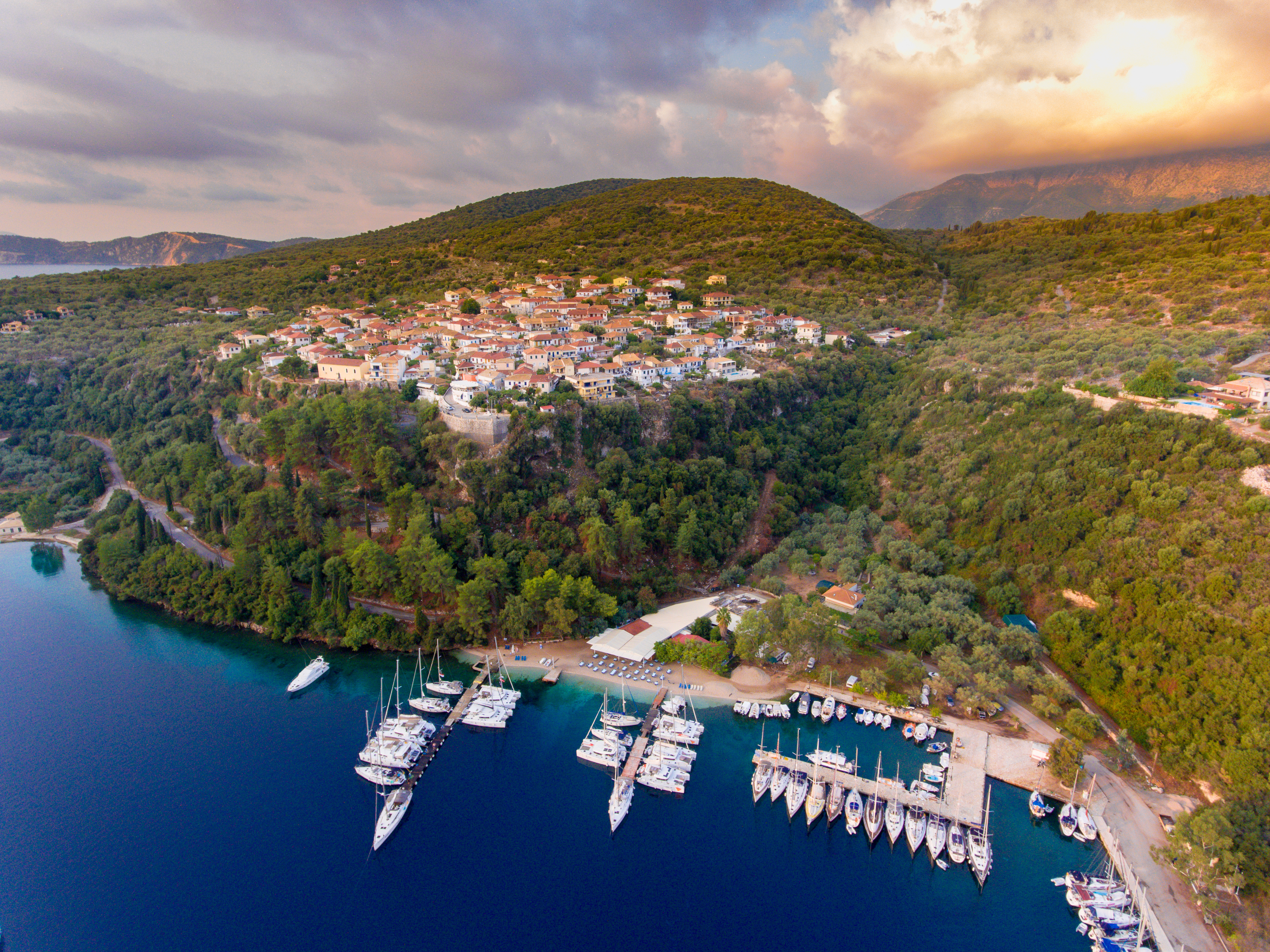 Aerial view of a Spartachori on Meganisi showing the moorings and pontoons. The coastal village is on a green hillside, with white houses and red roofs. Yachts dock at the marina beside calm, deep blue water—this could be your home for a fortnight south under a dramatic, cloud-filled sky at sunset.