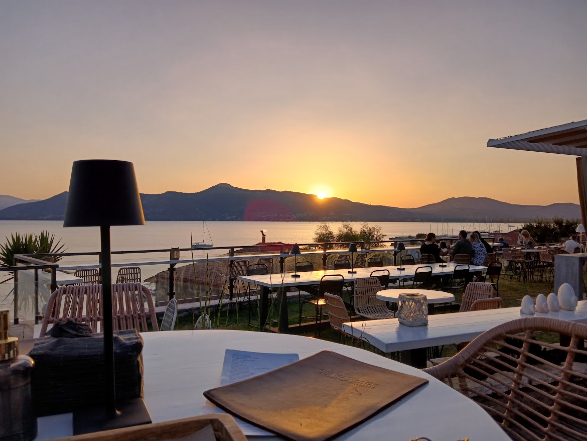 Outdoor restaurant terrace overlooking a calm sea at sunset, with distant mountains and sailboats—perfect for diners dreaming of a fortnight south. Tables and chairs are arranged under soft evening light, inviting you to savour the view.