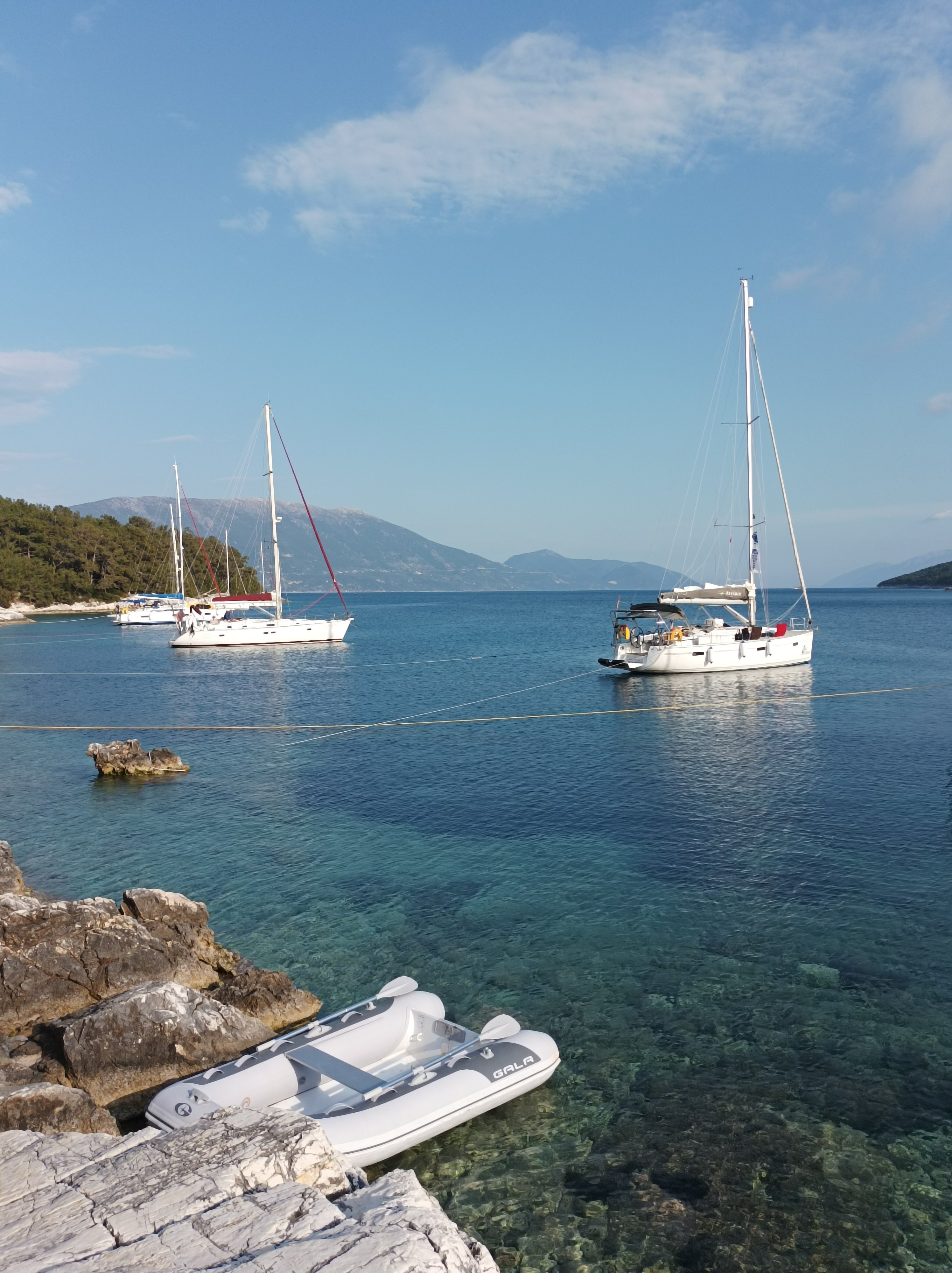 Port Atheni on Sail Ionians Quiet Escapes route. Two sailing boats are anchored in clear blue water near a rocky shoreline, with a small inflatable dinghy tied to the rocks—a tranquil stop along their route plan. Distant forested hills and mountains rise under a blue sky with scattered clouds.