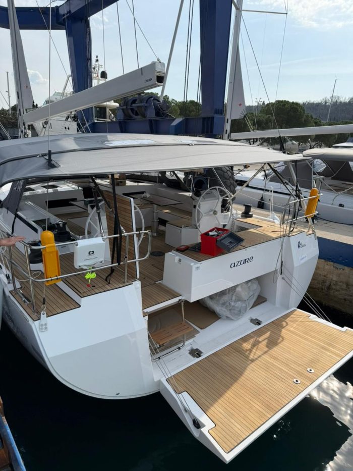 A modern white Bavaria C50 Greece named Azure is moored at a marina. This brand new yacht features a spacious wooden cockpit, dual steering wheels, and a lowered swim platform at the stern.