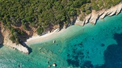 Aerial view of a secluded sandy beach with clear turquoise water, rocky cliffs, and dense green forest. A small boat is anchored near the shore—an idyllic spot to sail to Ithaca in the Ionian, where untouched beauty awaits discovery.