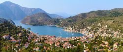 Panoramic view of a coastal village with red-roofed houses, green hills, and a blue bay dotted with anchored sailing boats under a clear sky—an inviting scene that captures the charm of the Ionian as you sail to Ithaca.