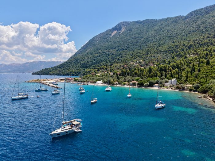 Sailing boats anchored in clear blue water near a small coastal village evoke the spirit of an Odyssey, as you embark on a memorable sailing holiday through lush green hills and a mountainous landscape under a partly cloudy sky—much like the legendary shores of Ithaca.