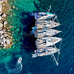 Four Sail Ionian yachts are moored side by side in clear turquoise water near rocks, viewed from above.
