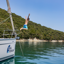 A person in blue swimming trunks jumps off a Sail Ionian boat into the clear Greek waters, with tree-covered hills in the background under a clear blue sky.
