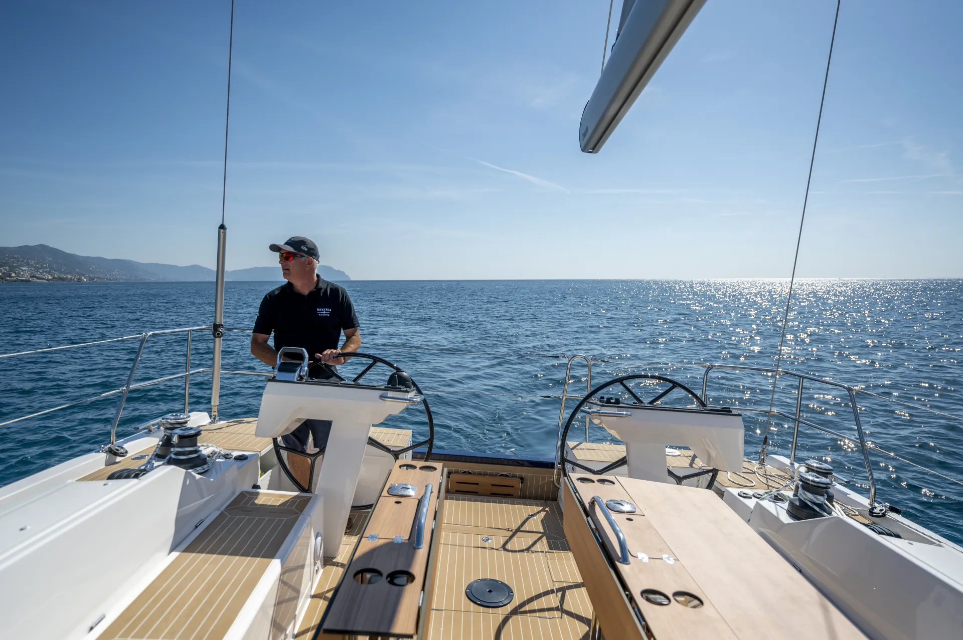 A person stands at the helm of a sleek Bavaria C46 sailboat on calm blue water under a clear sky, with distant land visible on the left.
