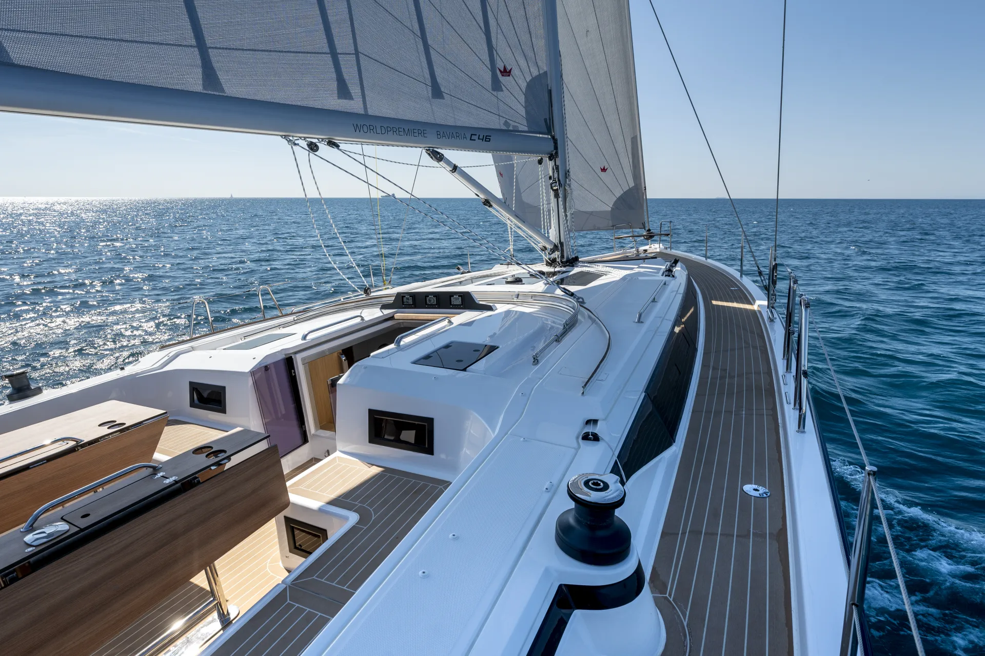 A modern Bavaria C46 sailboat with a raised white deck and wooden accents sails on calm blue water under a clear sky; its sails are up and sunlight reflects on the sea.
