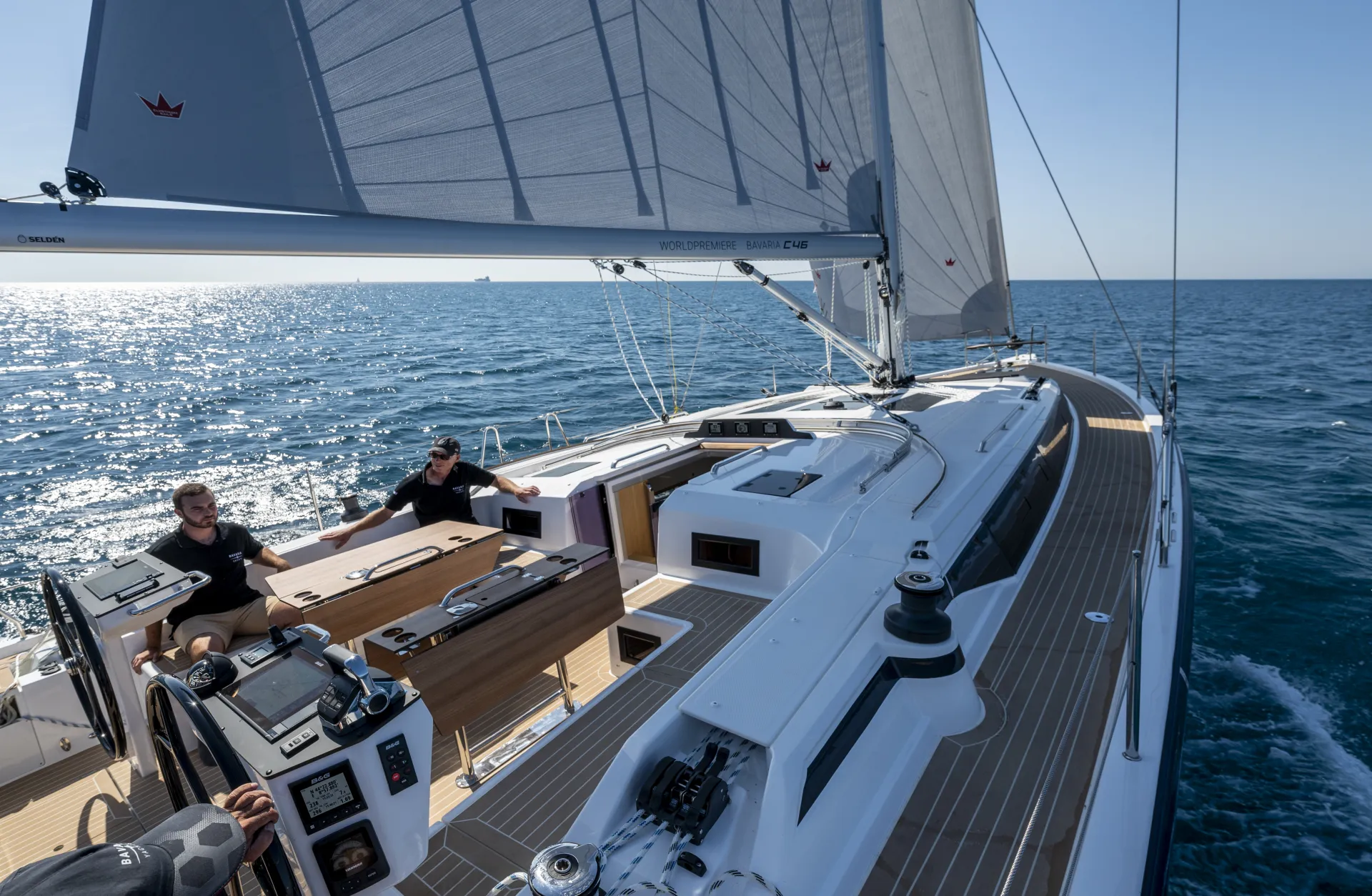 Two people sail a modern Bavaria C46 yacht on open water under a clear blue sky. The sun reflects off the calm sea, and the yacht’s deck features polished wood and advanced navigation instruments.