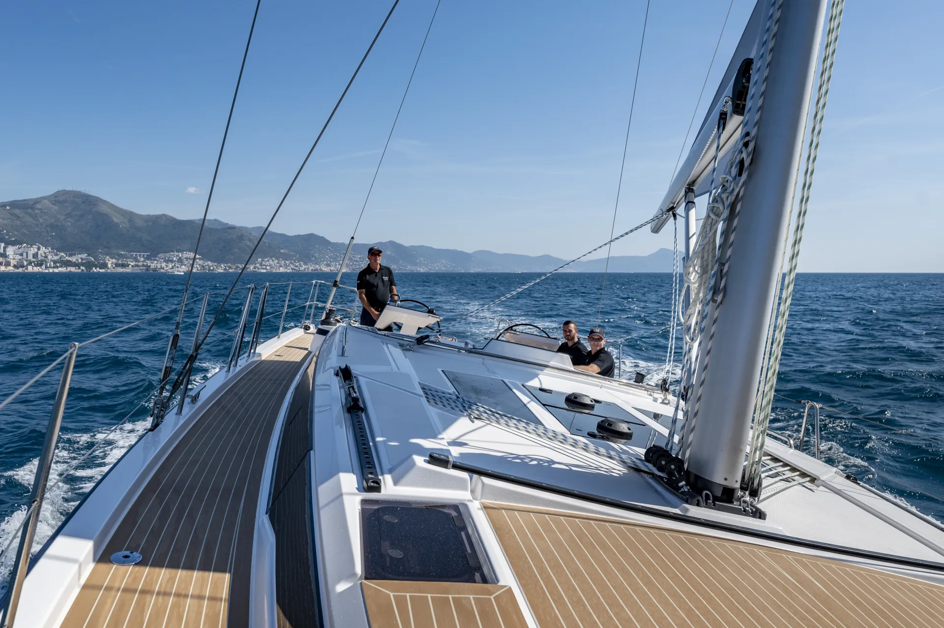 Three people are sailing a modern yacht on blue water with coastal mountains and a city visible in the background, under a clear sky.
