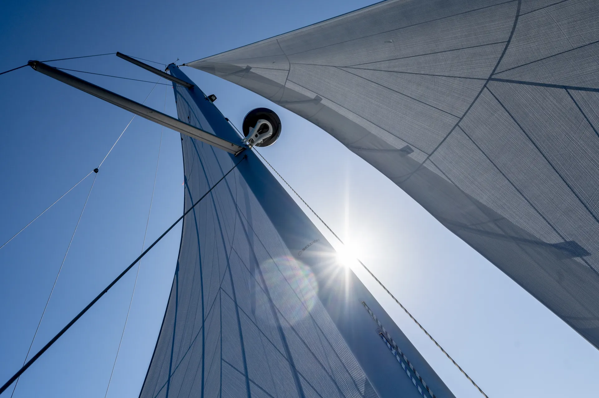 View from below of a sailboat’s sails against a clear blue sky, with sunlight shining through the sails and creating a lens flare. The image highlights the sail structure and the bright, sunny weather.