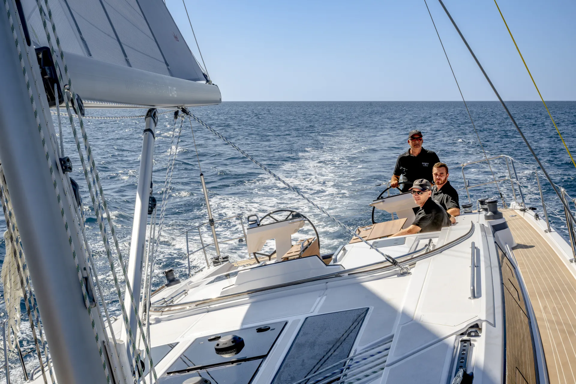 Three people are sailing on a white yacht in open water under a clear blue sky, with the sails up and wake in the sea behind them.