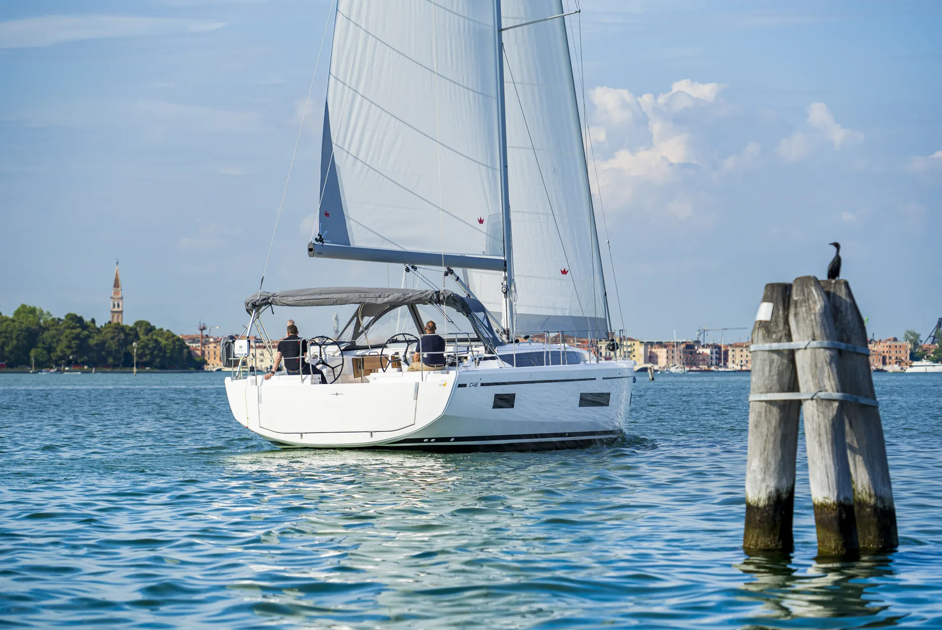 A white sailboat glides on calm blue water near a city shoreline, with a church tower visible in the background and a bird perched on a wooden post in the foreground.