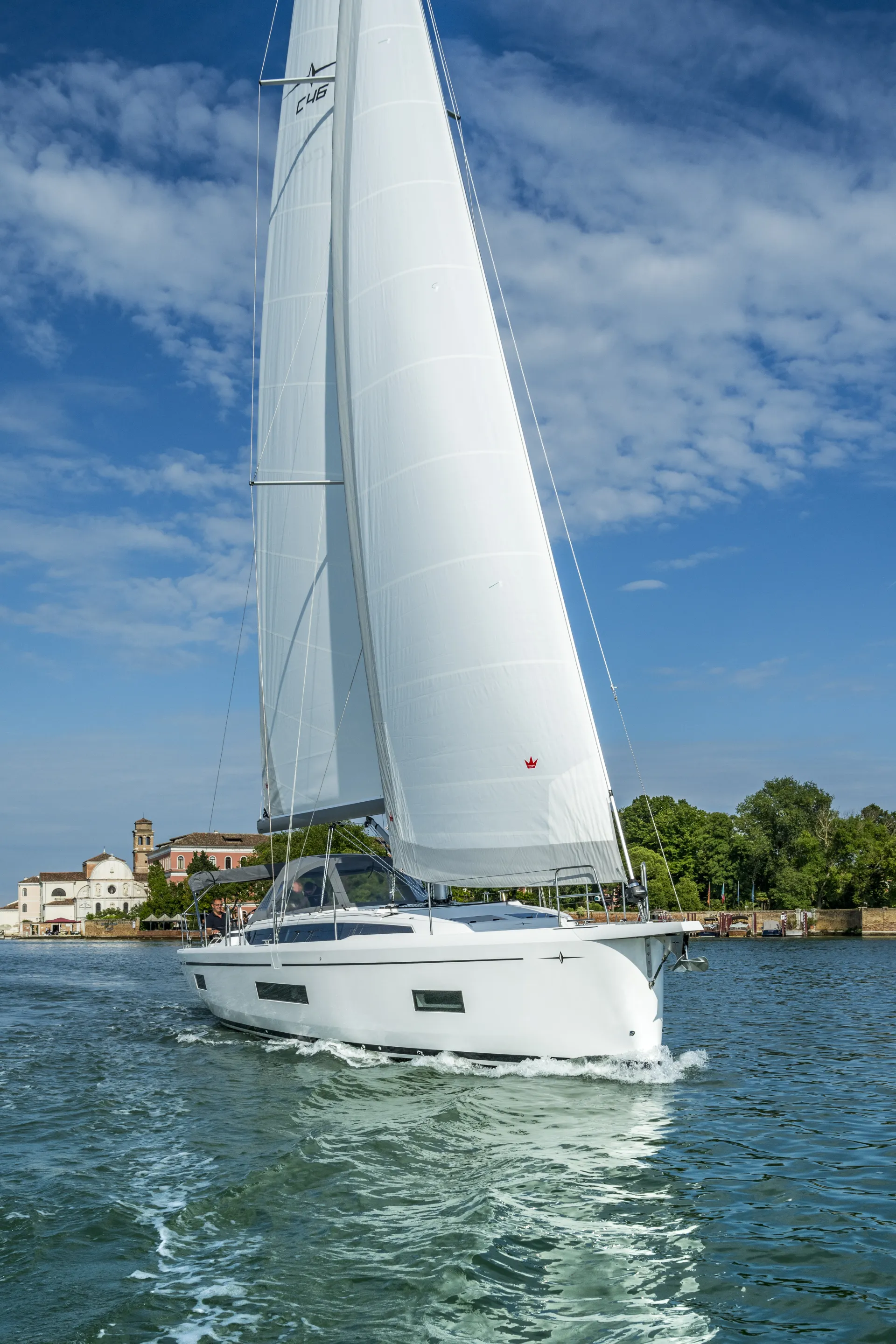 A white sailboat with raised sails glides on blue water near a shoreline with trees and buildings under a partly cloudy sky.