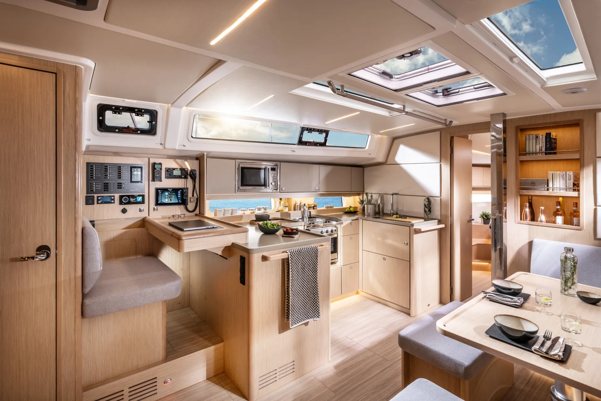 Modern yacht interior with a light wood finish, featuring a kitchen with appliances, a navigation area, dining table set for four, built-in shelves, and large windows letting in natural light.