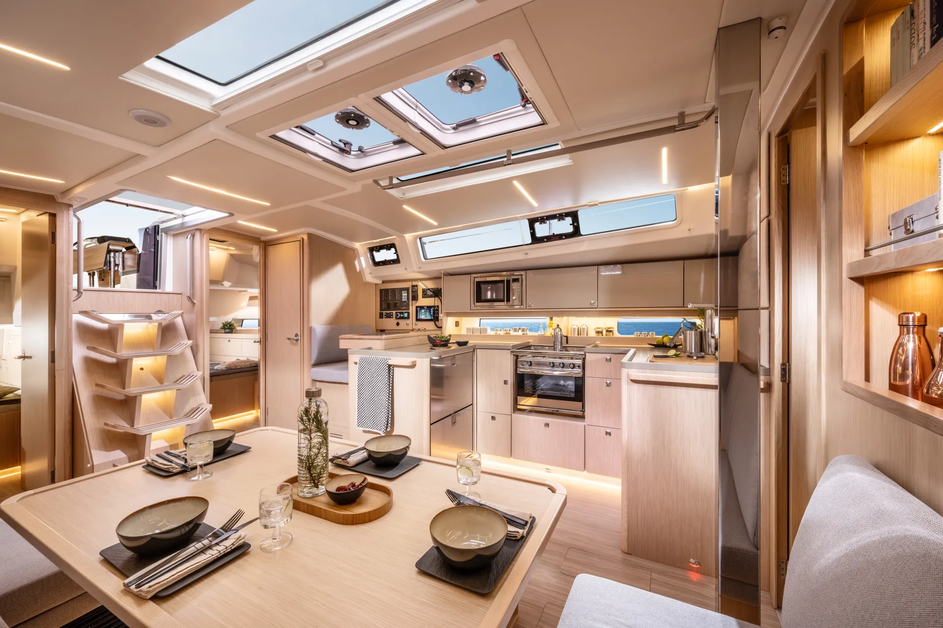 Modern yacht interior with light wood finishes, skylights, dining table set for four, kitchen area with hob and microwave, shelves with decor, and built-in lighting creating a bright, cosy atmosphere.