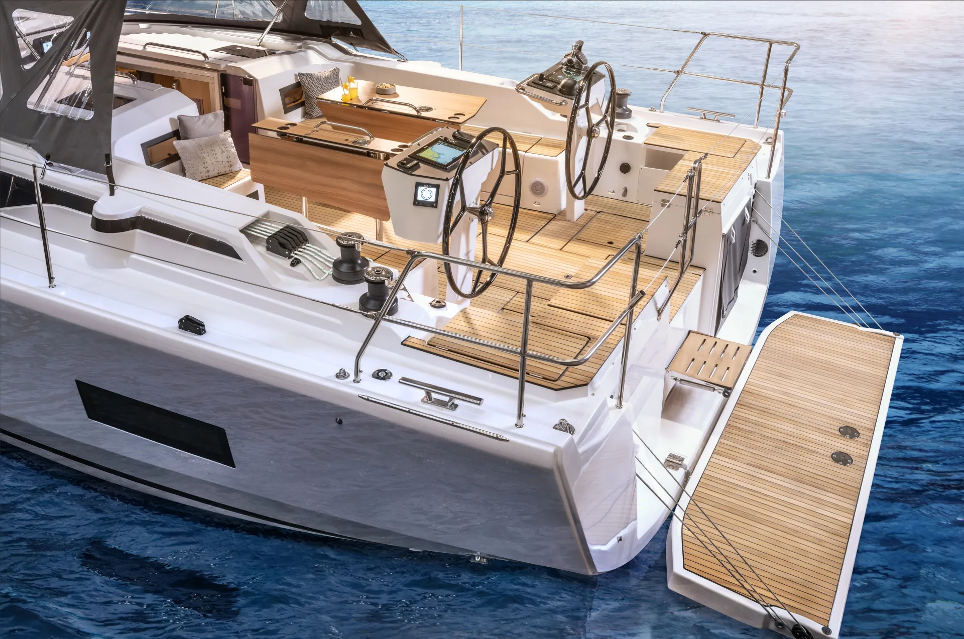 A modern sailboat with wooden decking, twin steering wheels, and an extended swim platform floats on clear blue water. The boat features cushioned seating and navigation controls.