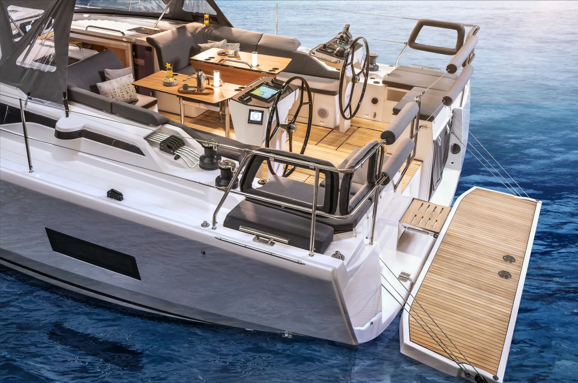 A modern sailboat’s aft deck with wooden flooring, a fold-down swim platform, two steering wheels, plush seating, a dining table with food and drinks, and control panels, floating on calm blue water.