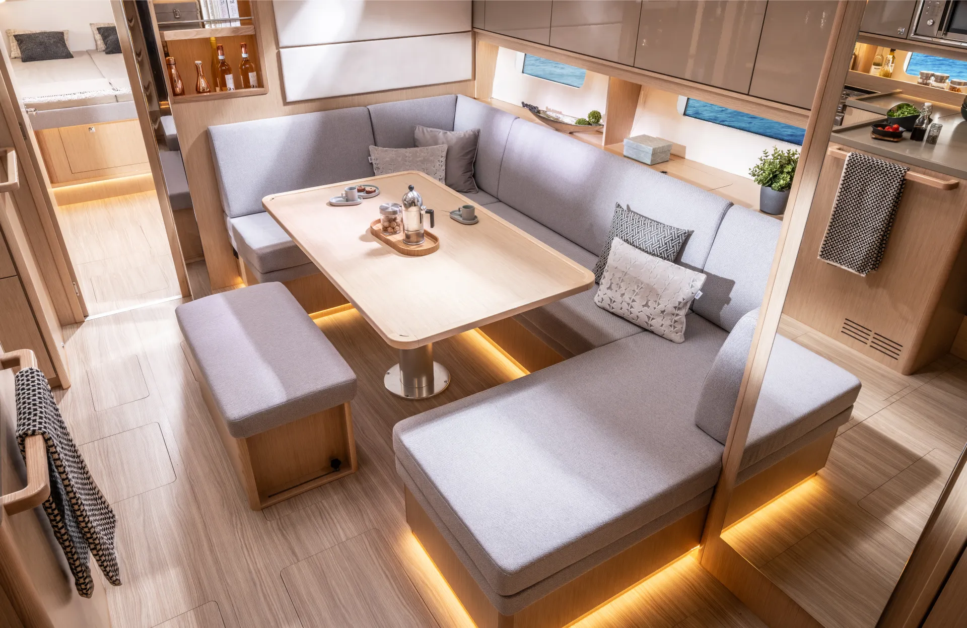 Modern yacht interior with light wood floors and cupboards, U-shaped grey seating around a rectangular table, soft lighting underneath benches, and windows showing sea views.