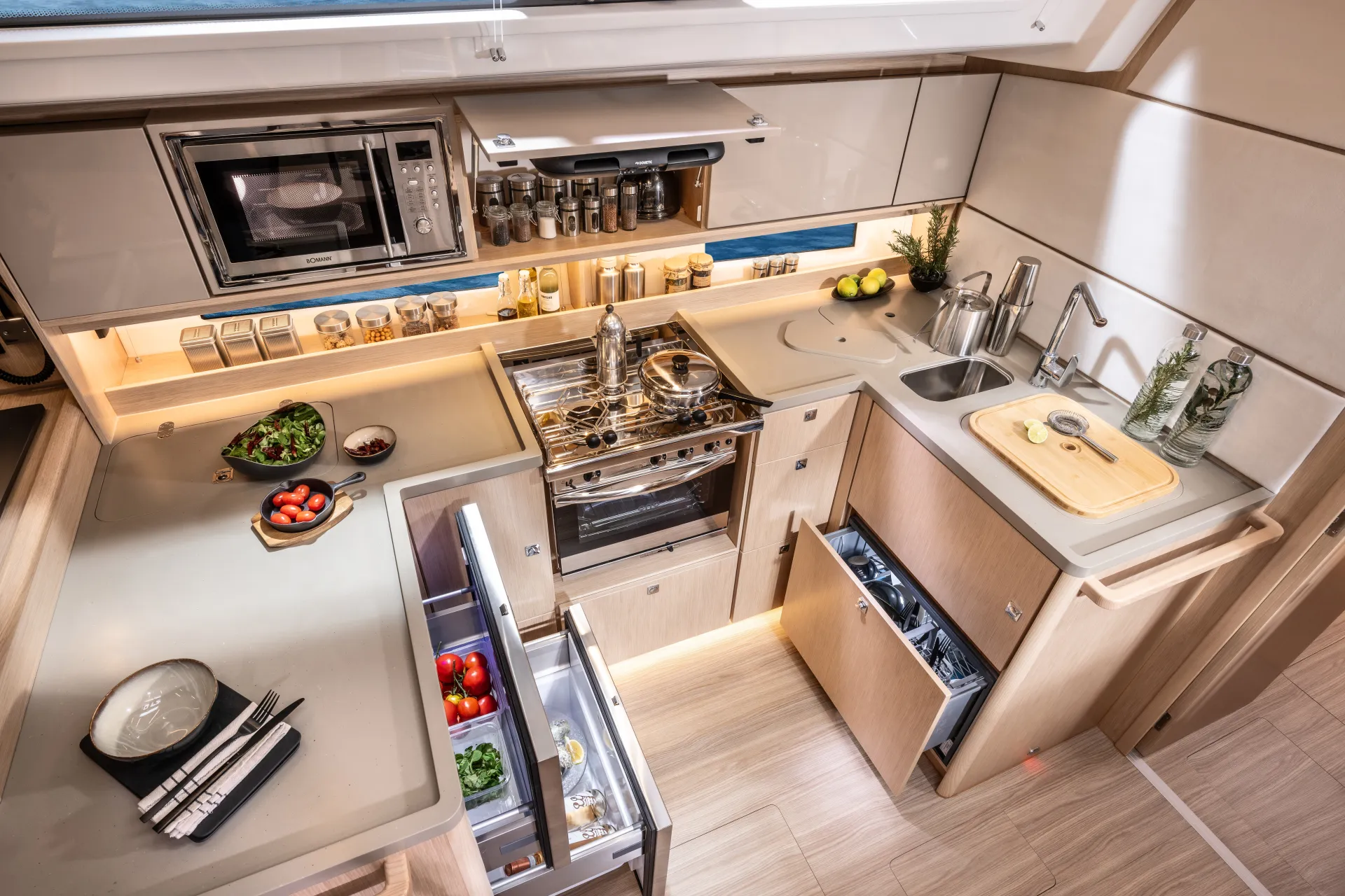 Modern yacht kitchen with open fridge, hob, microwave, sink, fresh ingredients, and utensils neatly arranged on light wood cupboards and worktops under bright lighting.