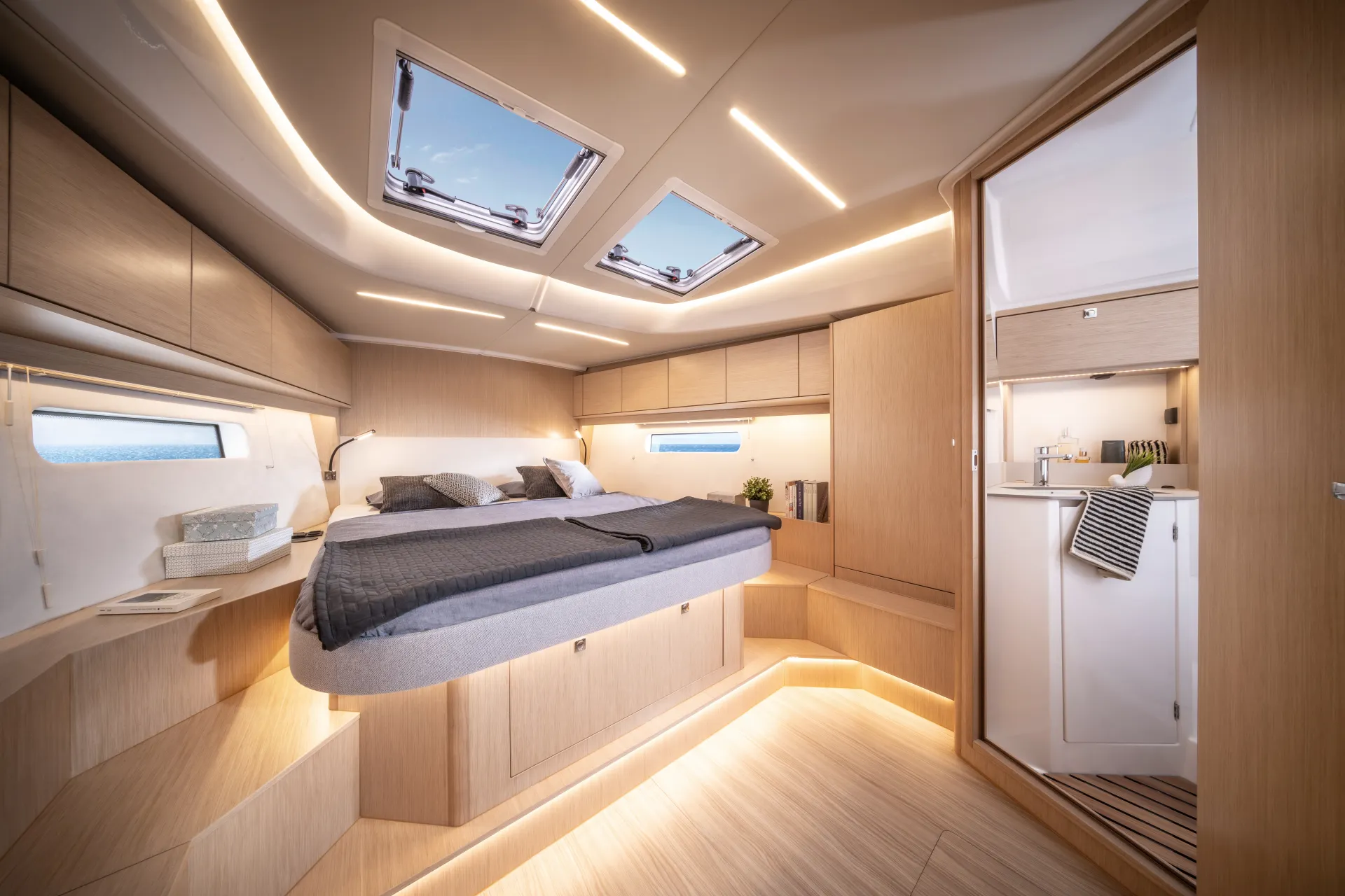 Modern yacht cabin with large bed, wood panelling, built-in cupboards, skylights, and an adjoining bathroom. Soft lighting highlights the clean, minimal design and natural light enters through windows and ceiling hatches.