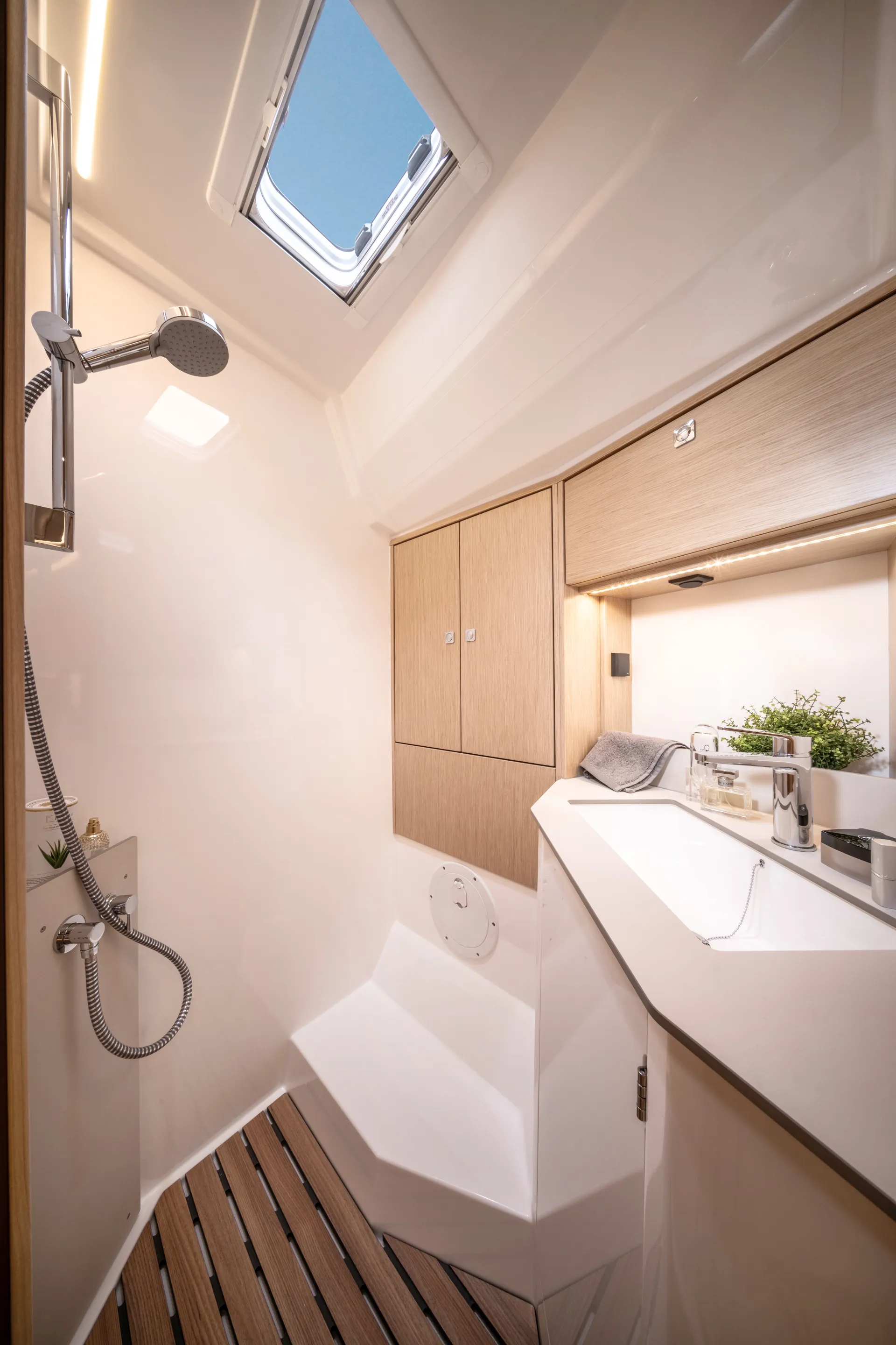Modern yacht bathroom with a skylight, wooden cupboards, a small basin, a shower, and wood-patterned flooring. A plant and towel sit near the basin, creating a bright, clean, and compact space.